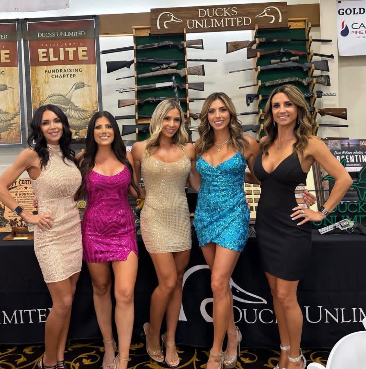 Five Mandi's Models in cocktail dresses at a Ducks Unlimited table with a rifle display