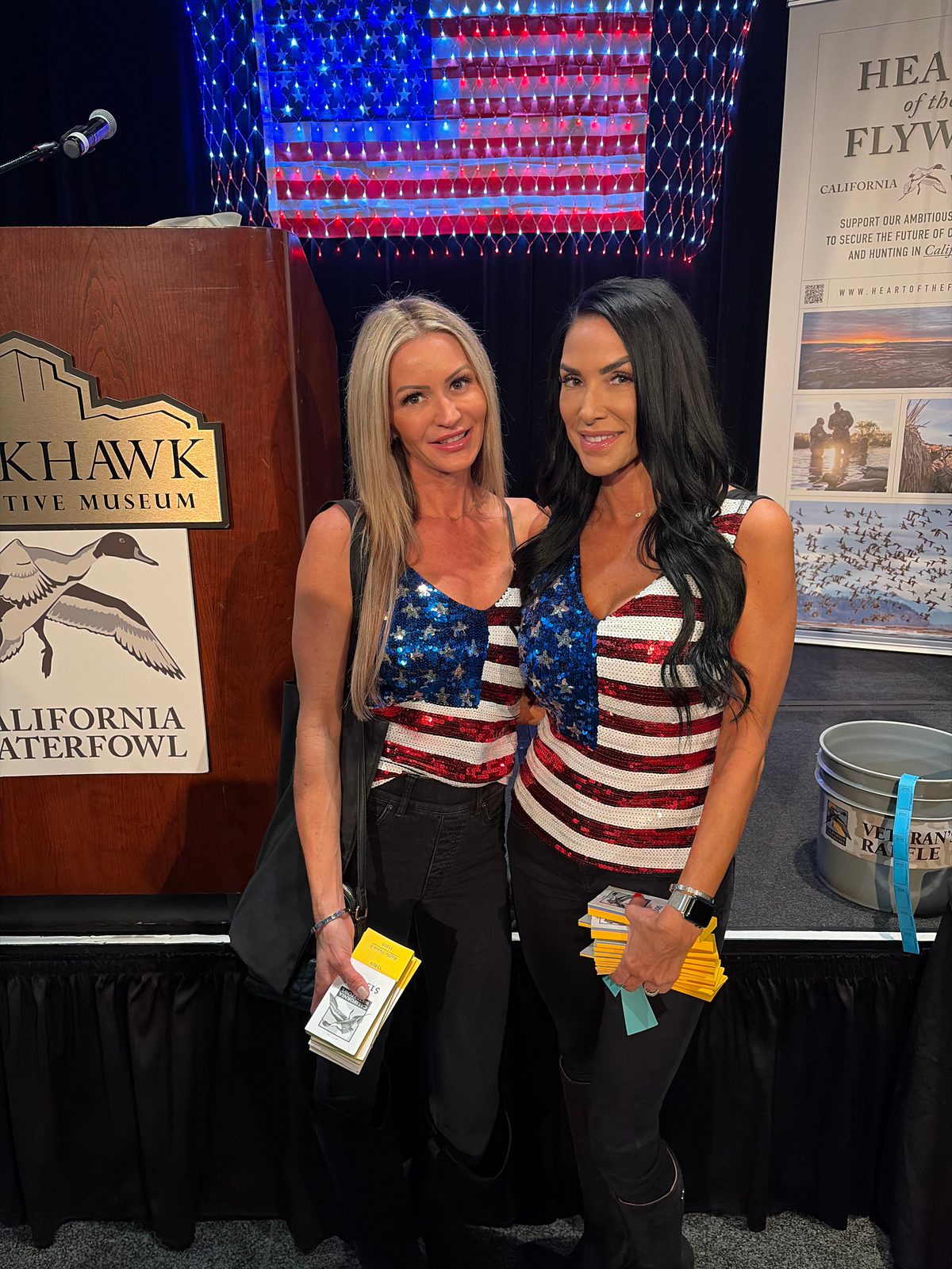 Two Mandi's Models in flag sequin tops holding raffle tickets beside a California Waterfowl podium
