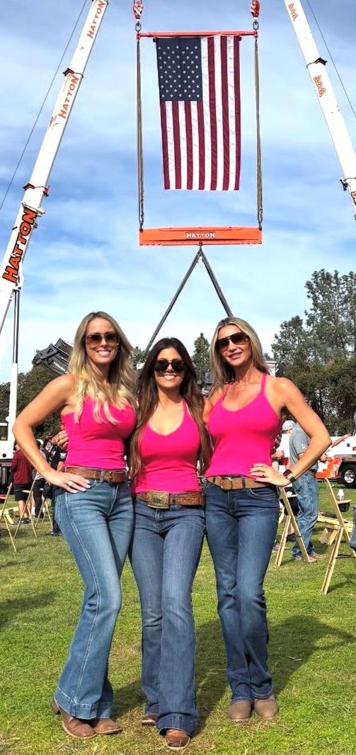 Three Mandi's Models in pink tanks and jeans outdoors under an American flag hung from cranes